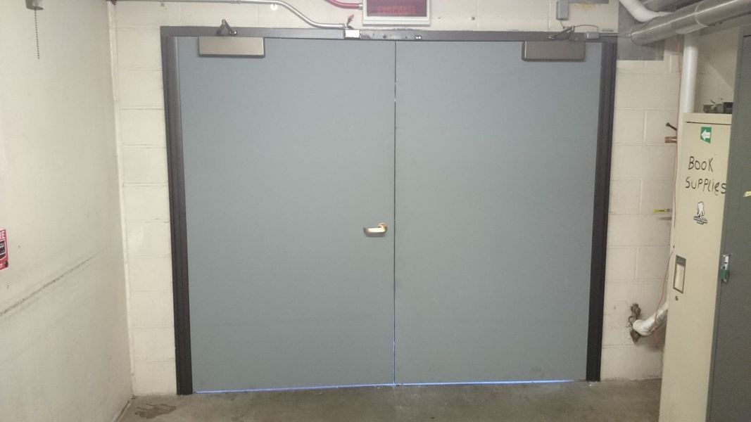 Gray double doors in a concrete hallway with door closers above.