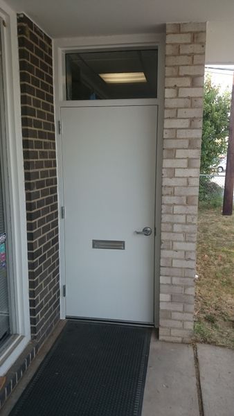 White door with mailbox and small window above, set in brick building. Black mat at entrance.