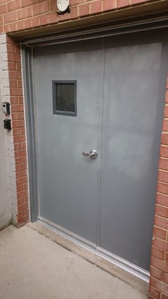 Gray metal door with small window, handle, and security panel in a brick building.