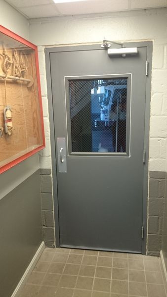 Gray metal door with window and hardware in a hallway, likely for security.