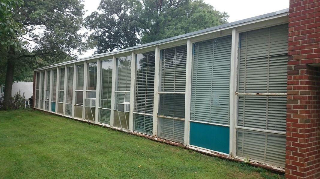 Long, glass-walled building with white frames and turquoise panels; brick wall on right, trees in background, and green grass.