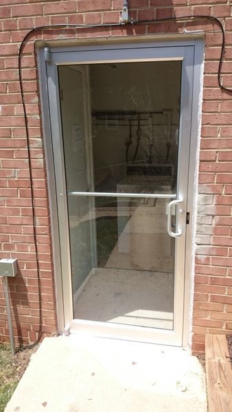 Clear glass door with silver frame, leading into a room with a white tank and other items visible. Set in a brick wall.