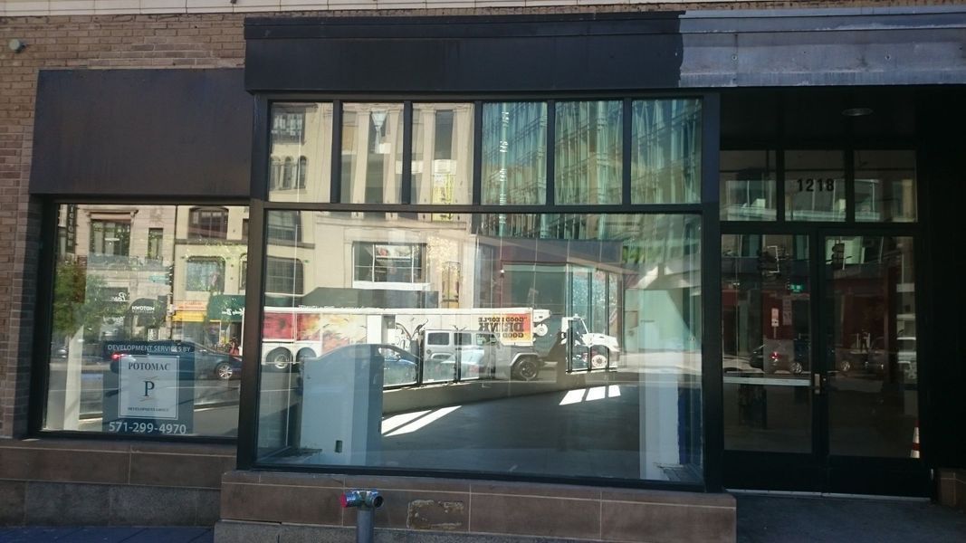 Storefront with large windows reflecting city buildings, a bus, and pedestrians. Black awning and door.