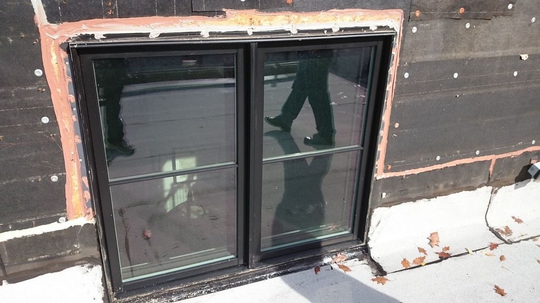Black framed window, set in dark wall. A person’s legs and feet are reflected in the glass.