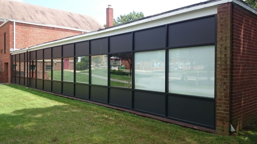 Long, black-framed windows along a brick building. Some windows have blinds, and the grass is green.