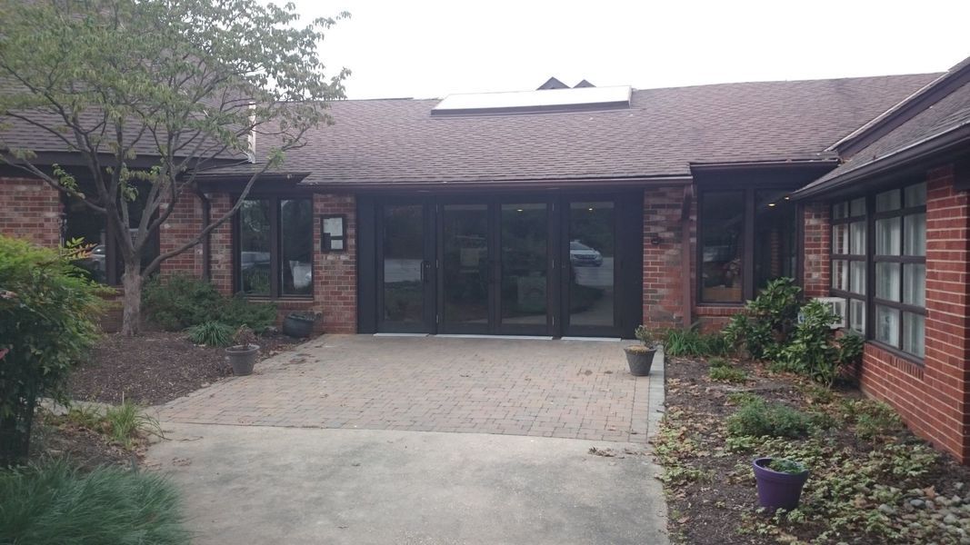 Brick building with glass doors and windows, a concrete walkway, and surrounding greenery.