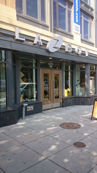 La-Z-Boy store front with glass doors and windows, beige exterior, and sidewalk.
