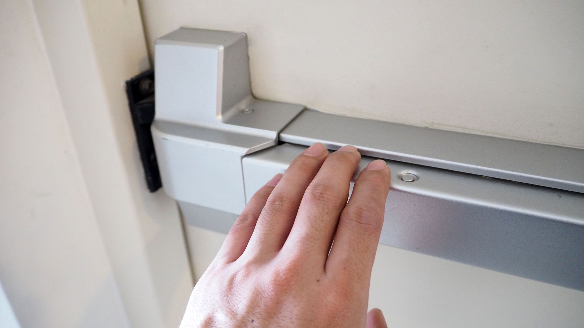 Hand reaching for a silver, metal push bar on a cream-colored door.