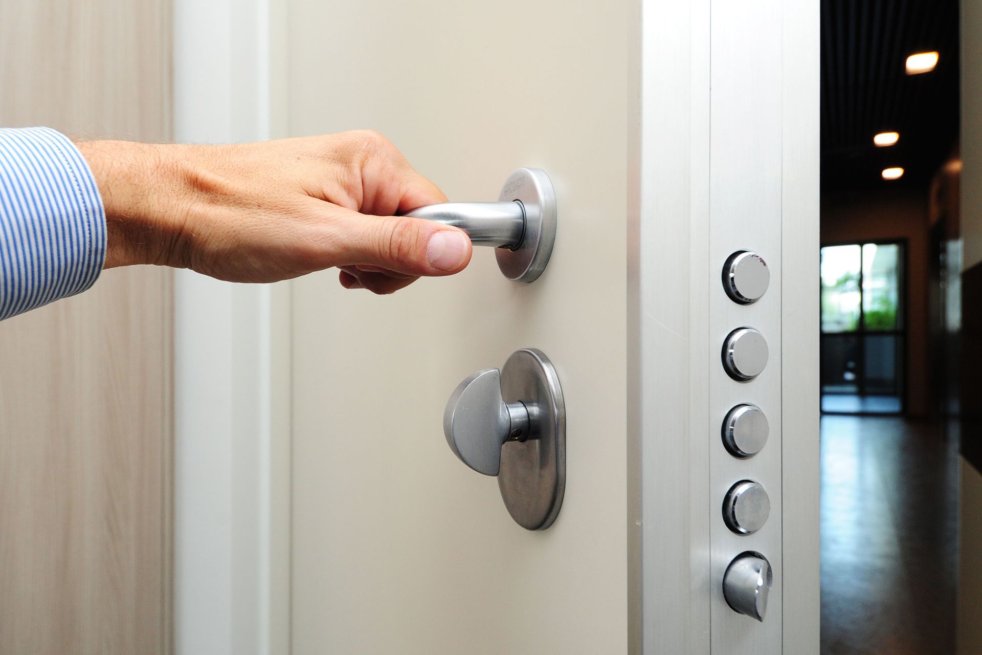 Person's hand on a silver door handle, opening a white door. A keypad is visible on the side.