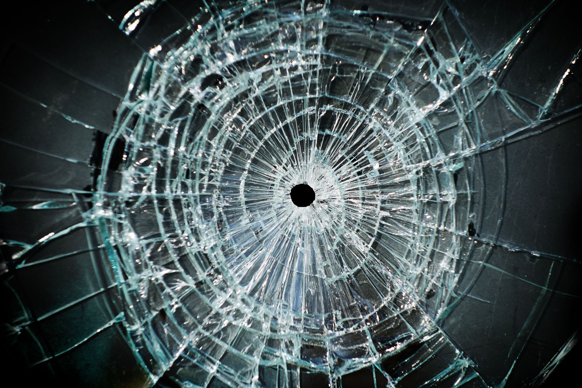 Shattered glass with a central hole, spiderweb-like cracks radiate outwards against a dark background.