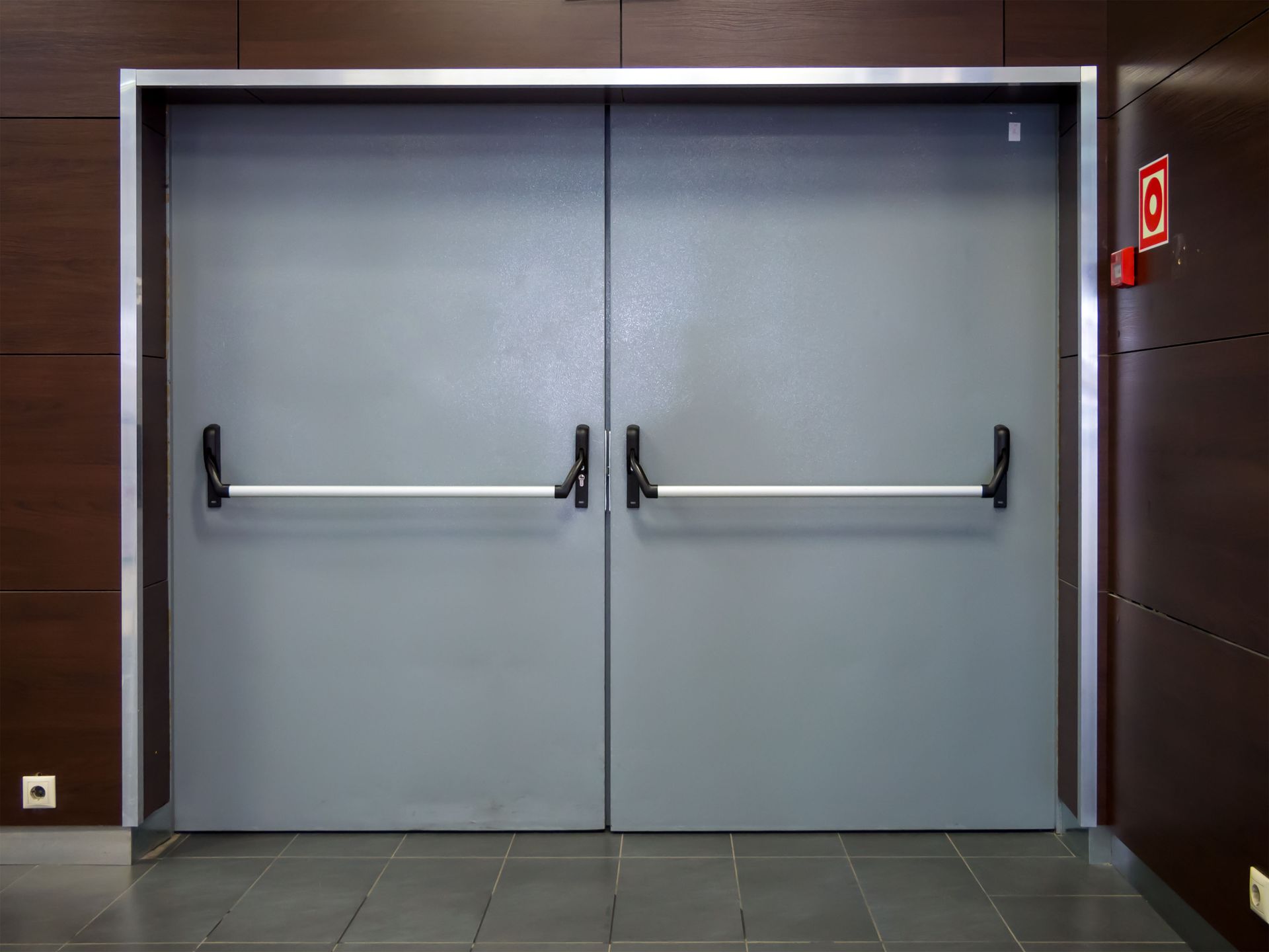 Double gray fire exit doors with push bars, silver frame, and fire alarm sign.