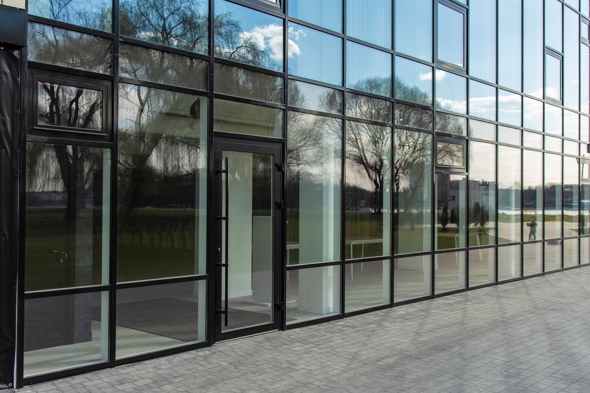 Modern building with glass windows reflecting trees and sky; black frames and entrance door.