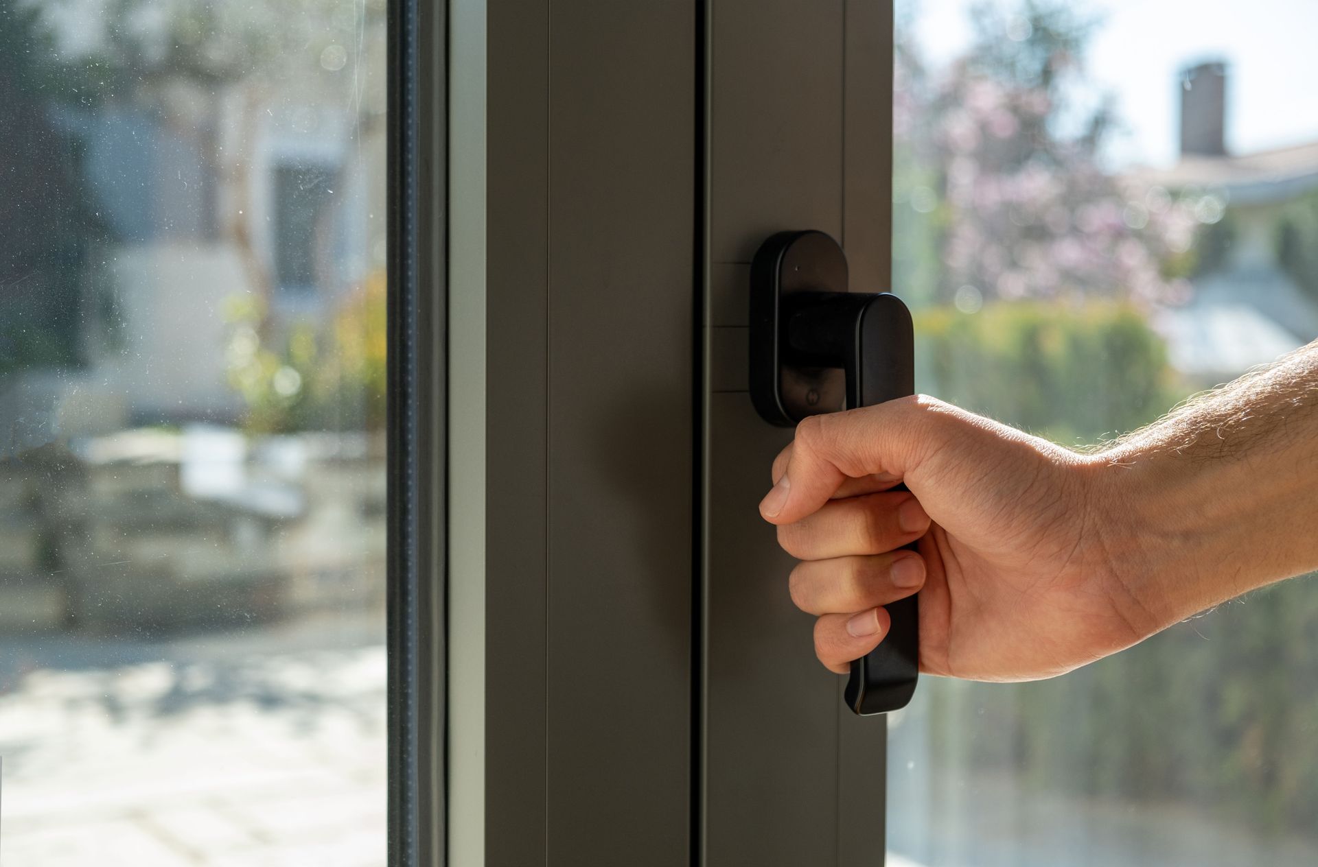 Hand holding a black window handle, outdoors. Gray window frame, visible garden and house in background.