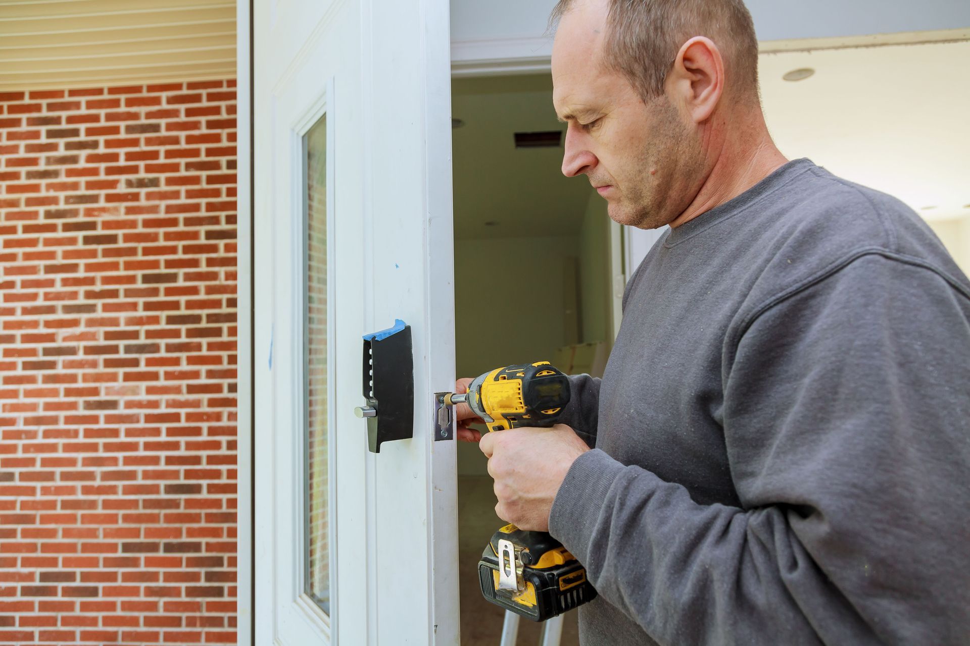 Man installing a smart lock on a white door with a drill outside a brick building.