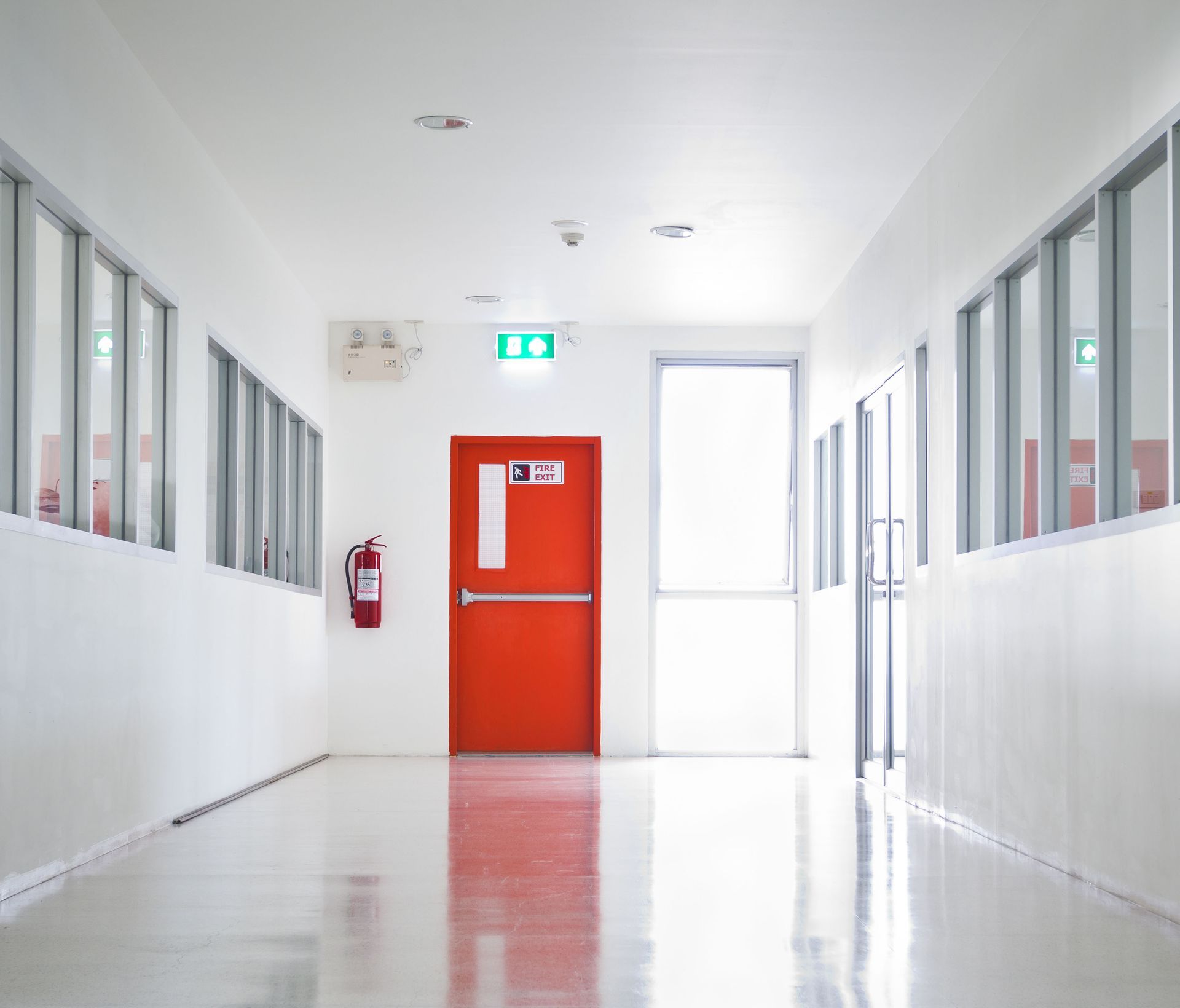 Bright hallway with red emergency door and fire extinguisher. Windows line walls. Exit sign visible.