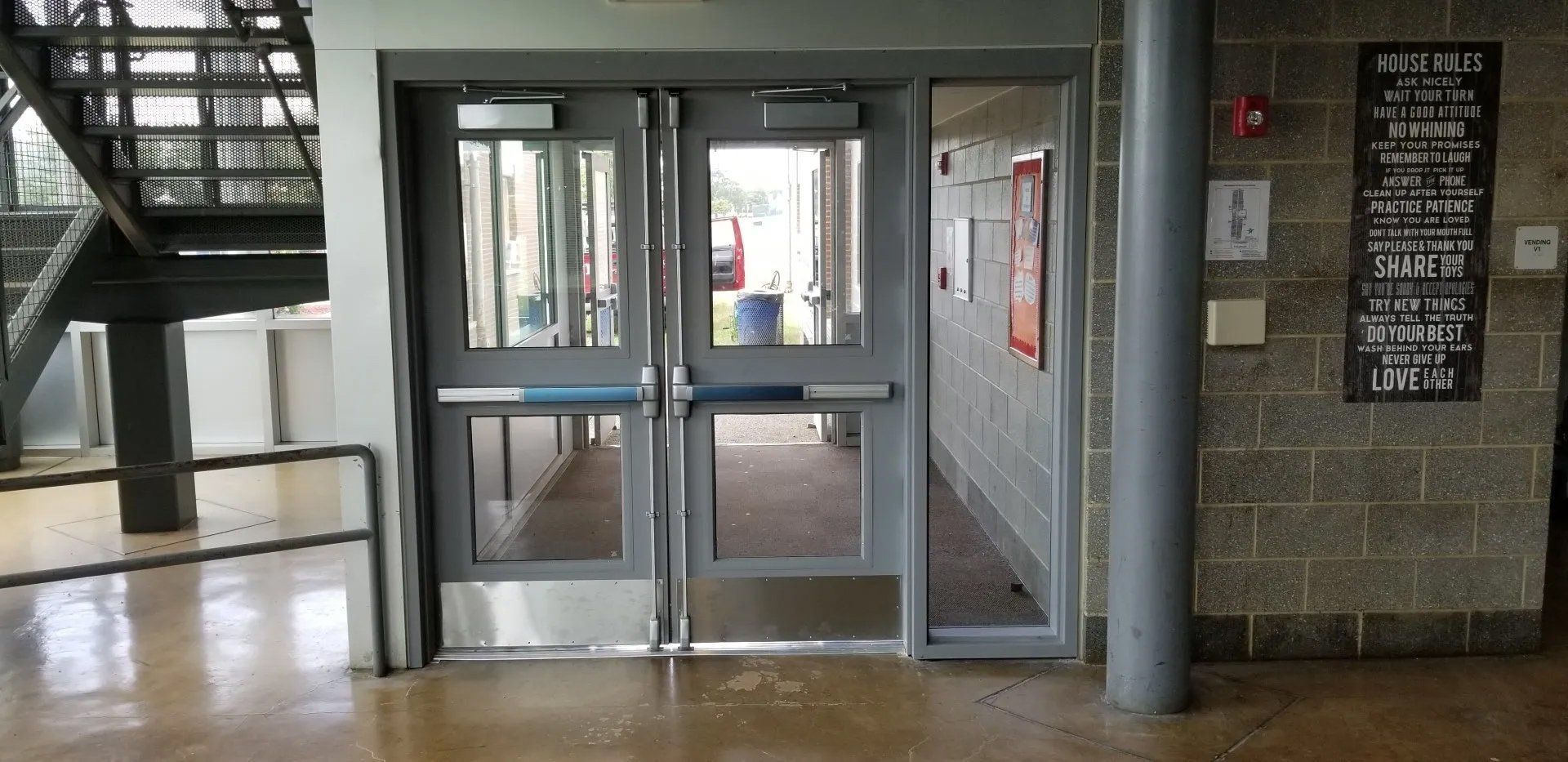 Double doors with windows open to a hallway. Signage and a fire alarm are mounted on the brick wall to the right.