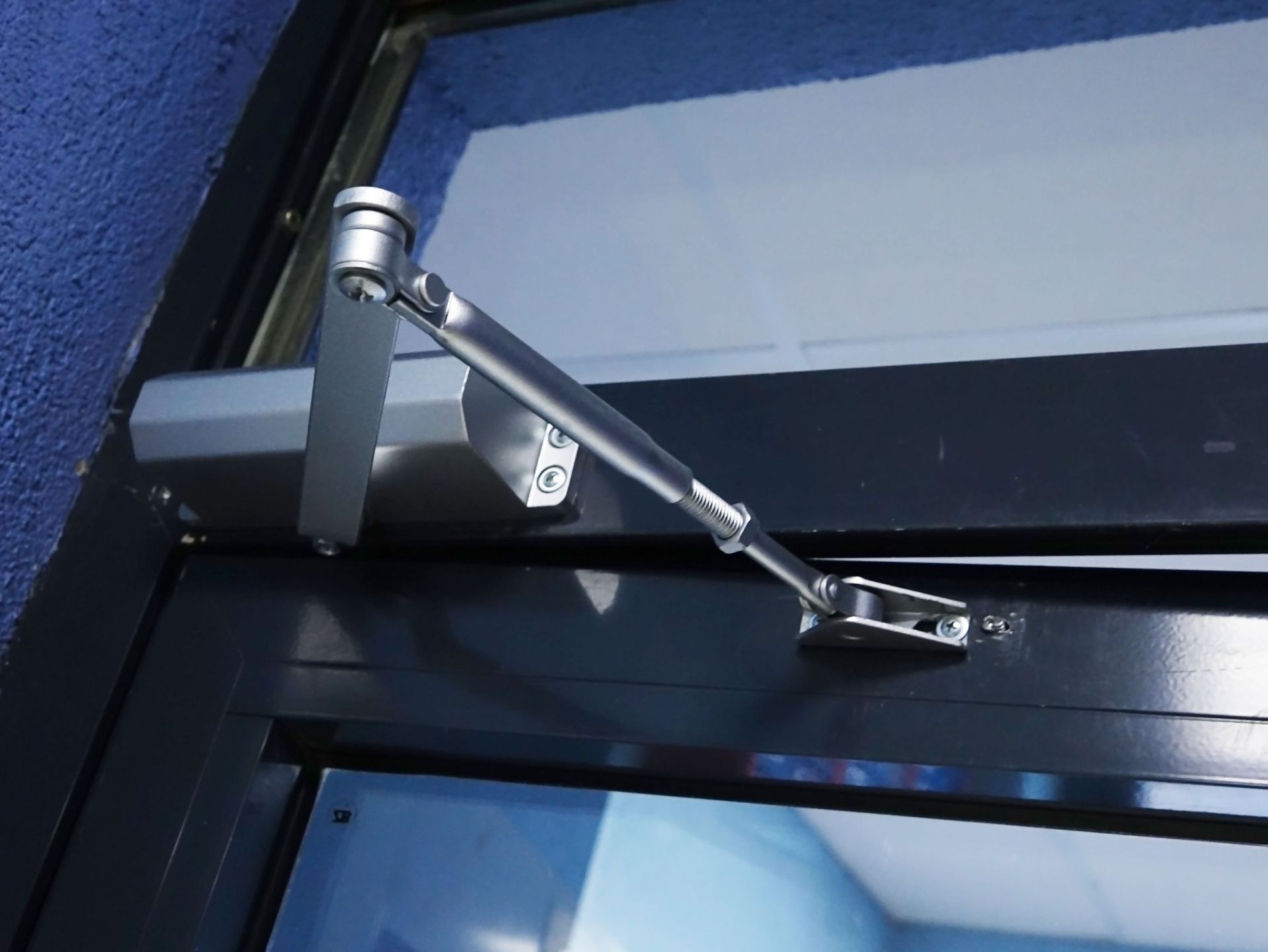 Door closer on a dark door frame. Silver arm and hydraulic cylinder.