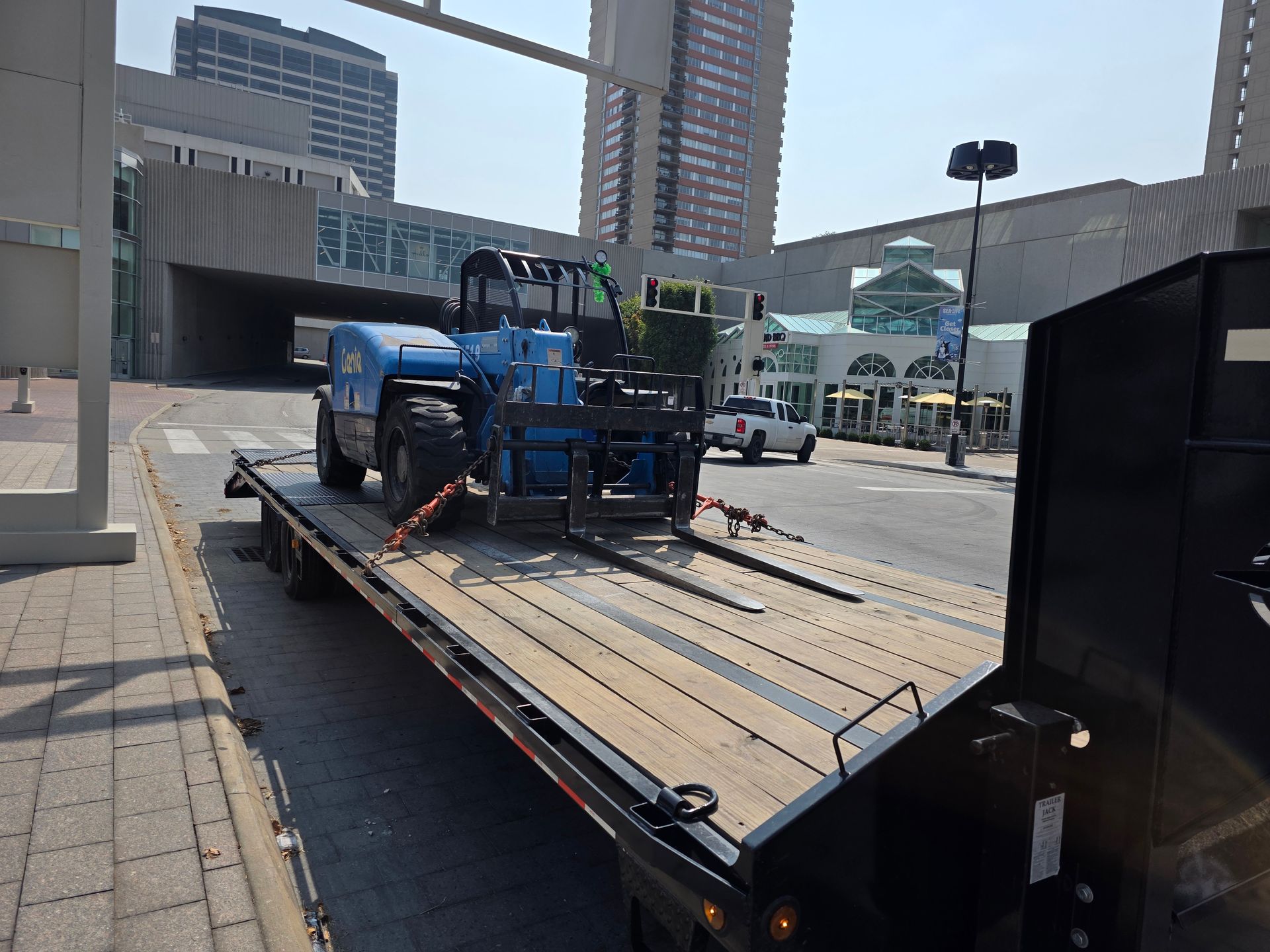Blue forklift on a flatbed trailer, street setting. Buildings in the background. Sunny day.