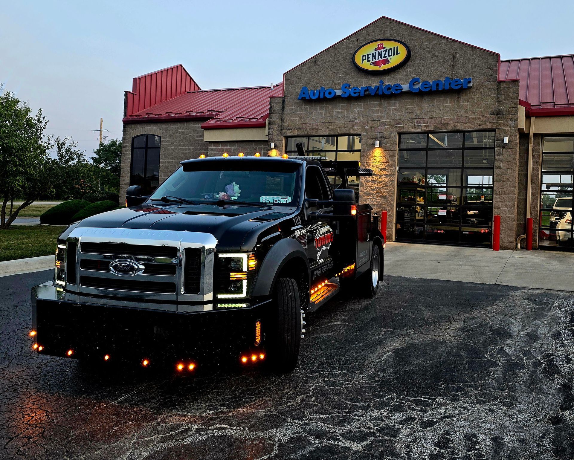 Black tow truck parked in front of a car repair shop with red roof, under a dusky sky.