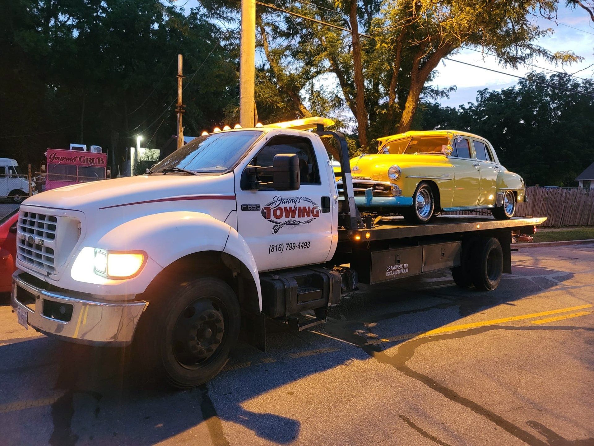 Tow truck carrying a yellow vintage car on a flatbed, parked on a street at dusk.