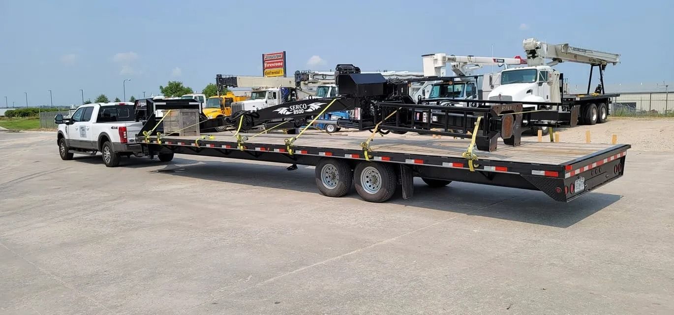 A white pickup truck towing a trailer with construction equipment on a sunny day.