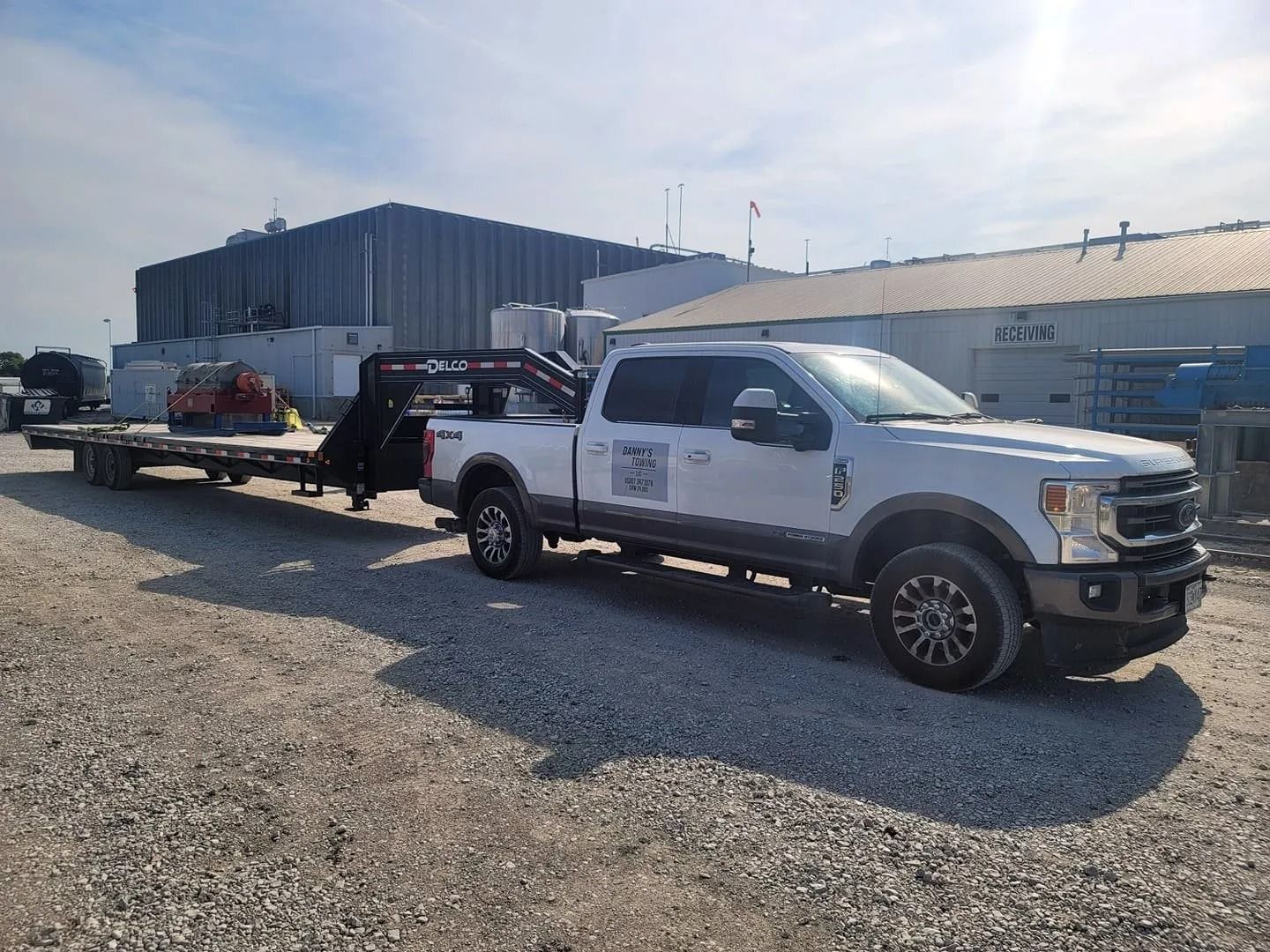 White pickup truck hauling a flatbed trailer on gravel in front of industrial buildings.