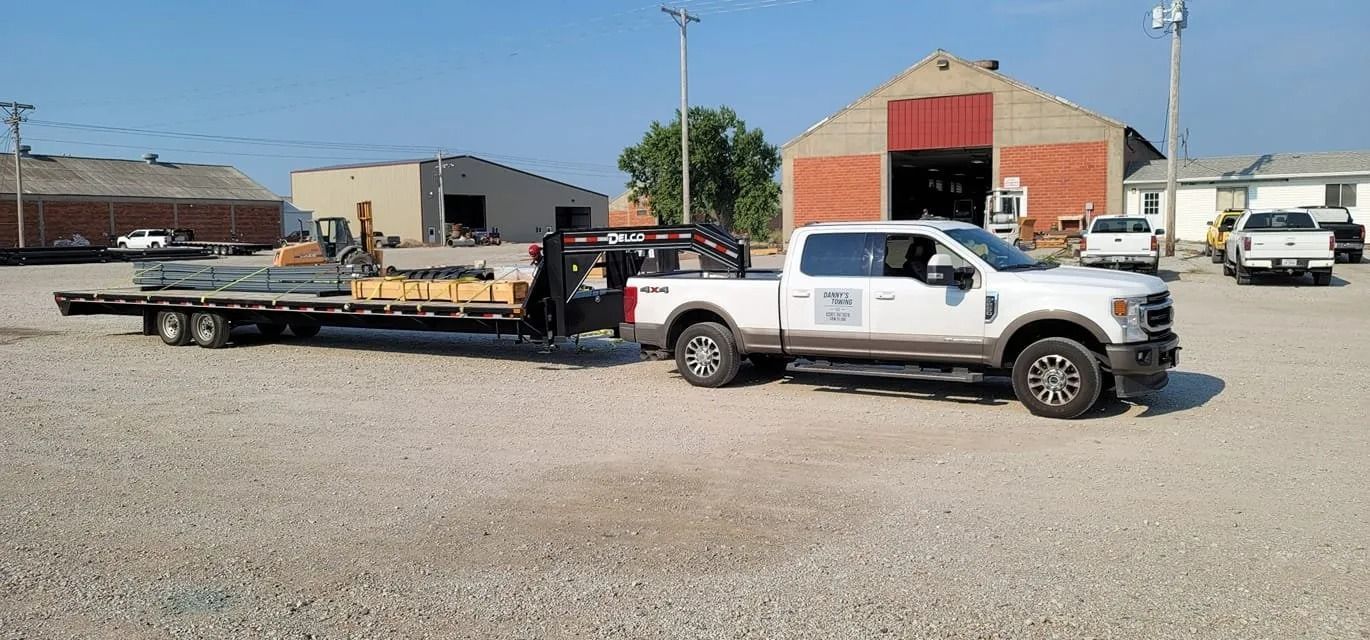 White pickup truck towing a flatbed trailer with materials. Parked on gravel near buildings and a barn.