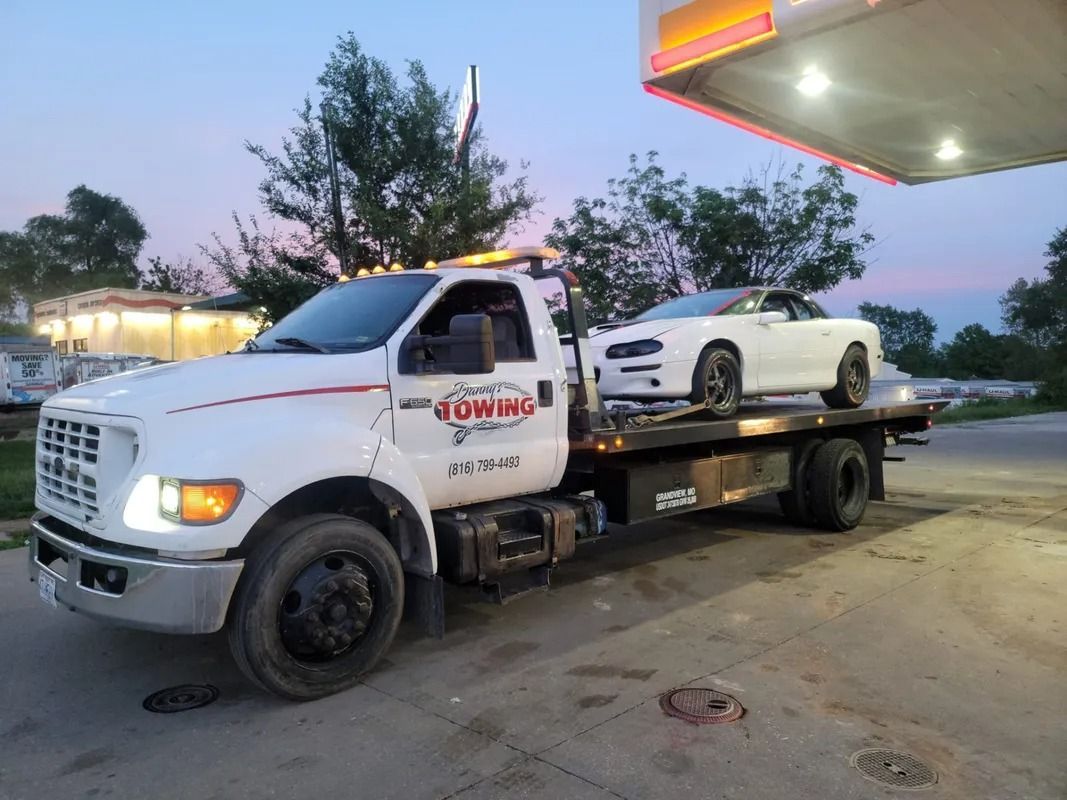 White tow truck carrying a white sports car at a gas station at dusk.