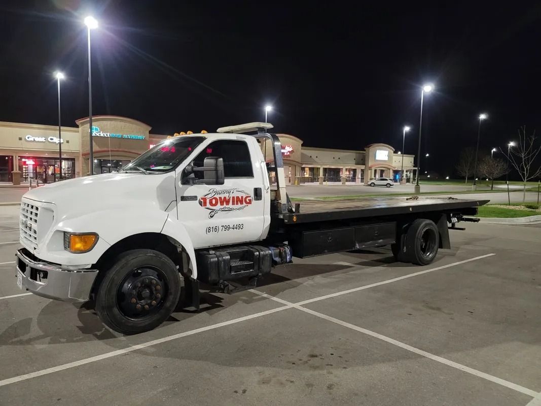 White tow truck parked at night in a parking lot with storefronts in the background.
