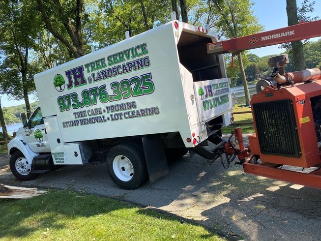 White tree service truck and stump grinder on a paved driveway near water.