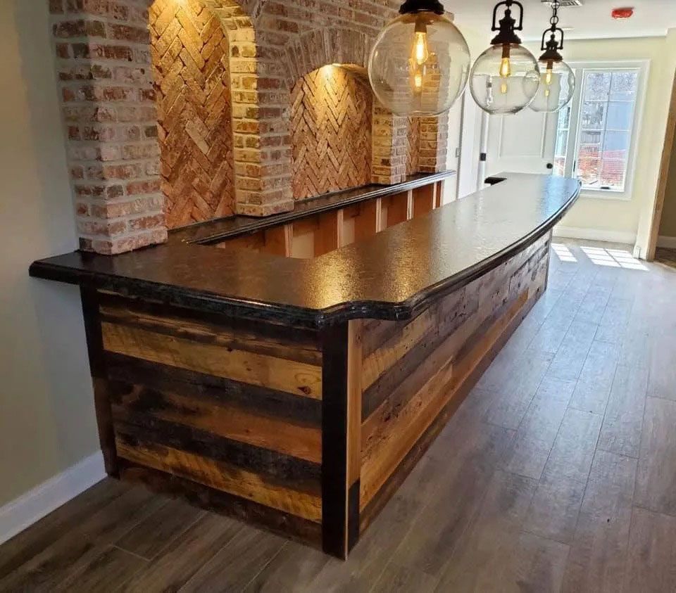 Rustic wooden bar counter with black countertop in a bright brick-walled room, lit by hanging pendant lights