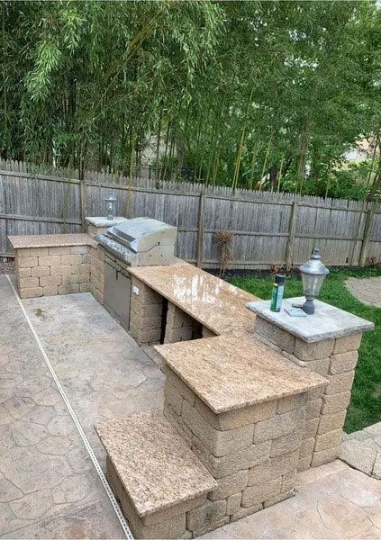Outdoor stone patio with built-in barbecue, benches, and a small table beside a lawn and bamboo fence