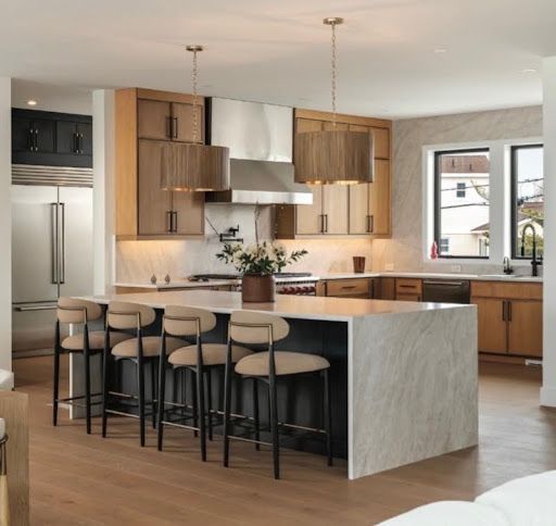 Modern kitchen with marble island, black stools, wood cabinets, and stainless steel appliances.