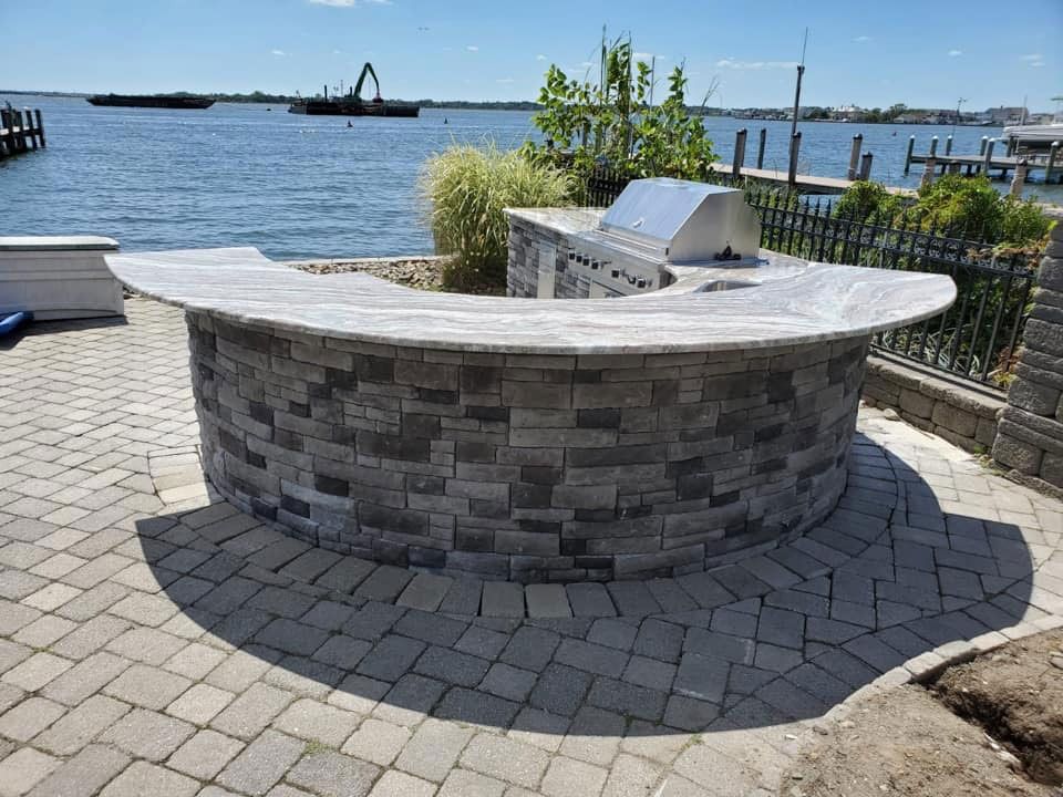Curved stone seating area beside a waterfront with pavers, railing, and boats in the distance