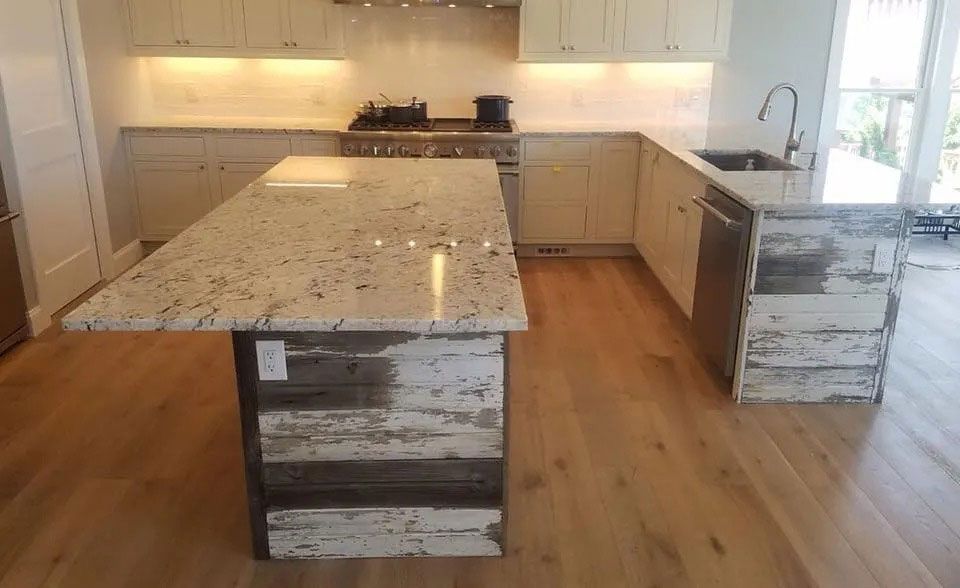 Modern kitchen with a large granite island, white cabinets, hardwood floors, and a windowed sink area.