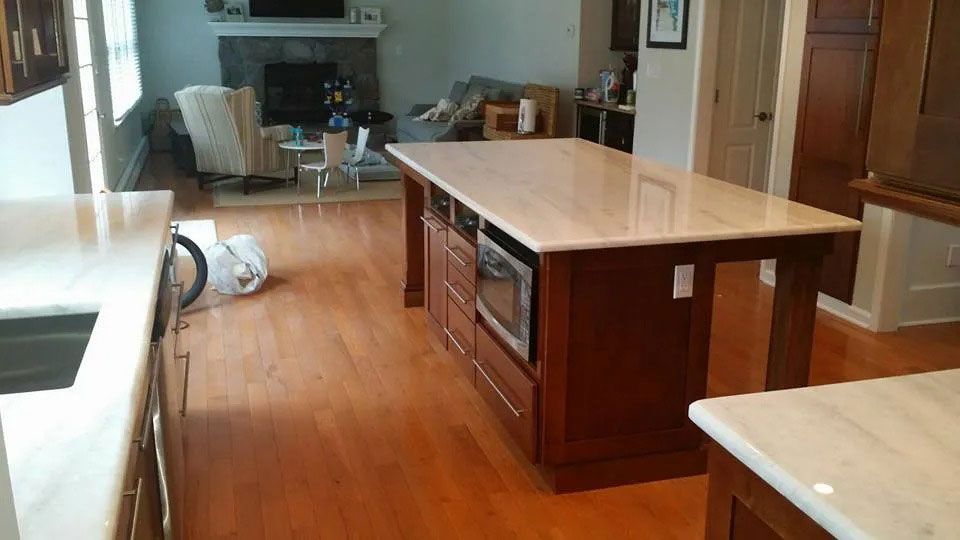 Modern kitchen with a large island, hardwood floors, and light countertops; living area visible in the background
