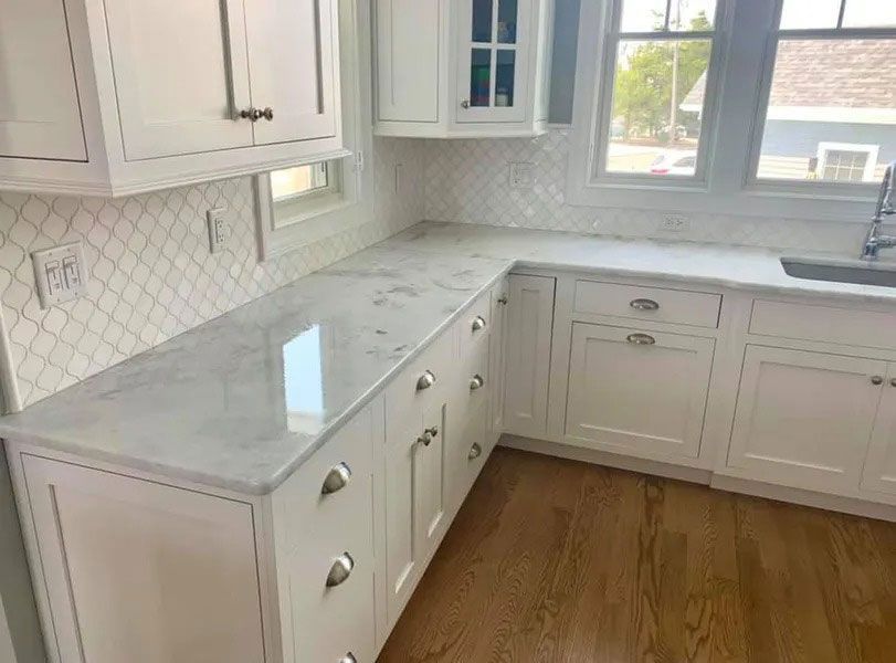 Bright white kitchen with marble countertops, white cabinets, and a window over wood floors