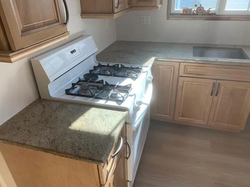 Small kitchen with white gas stove, granite counter, wood cabinets, and a sink by the window.