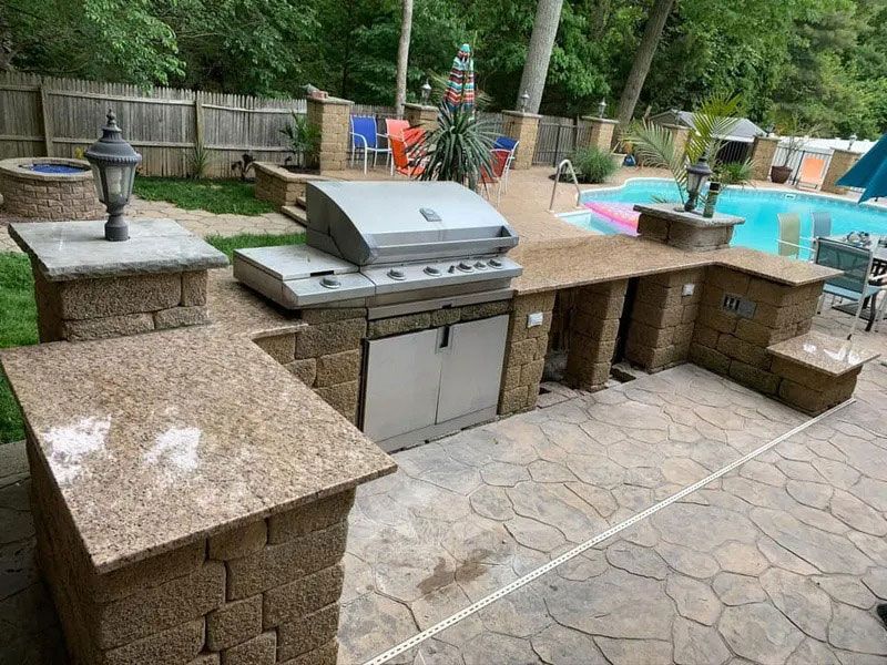 Backyard outdoor kitchen with stone counters, built-in grill, and patio by a swimming pool.