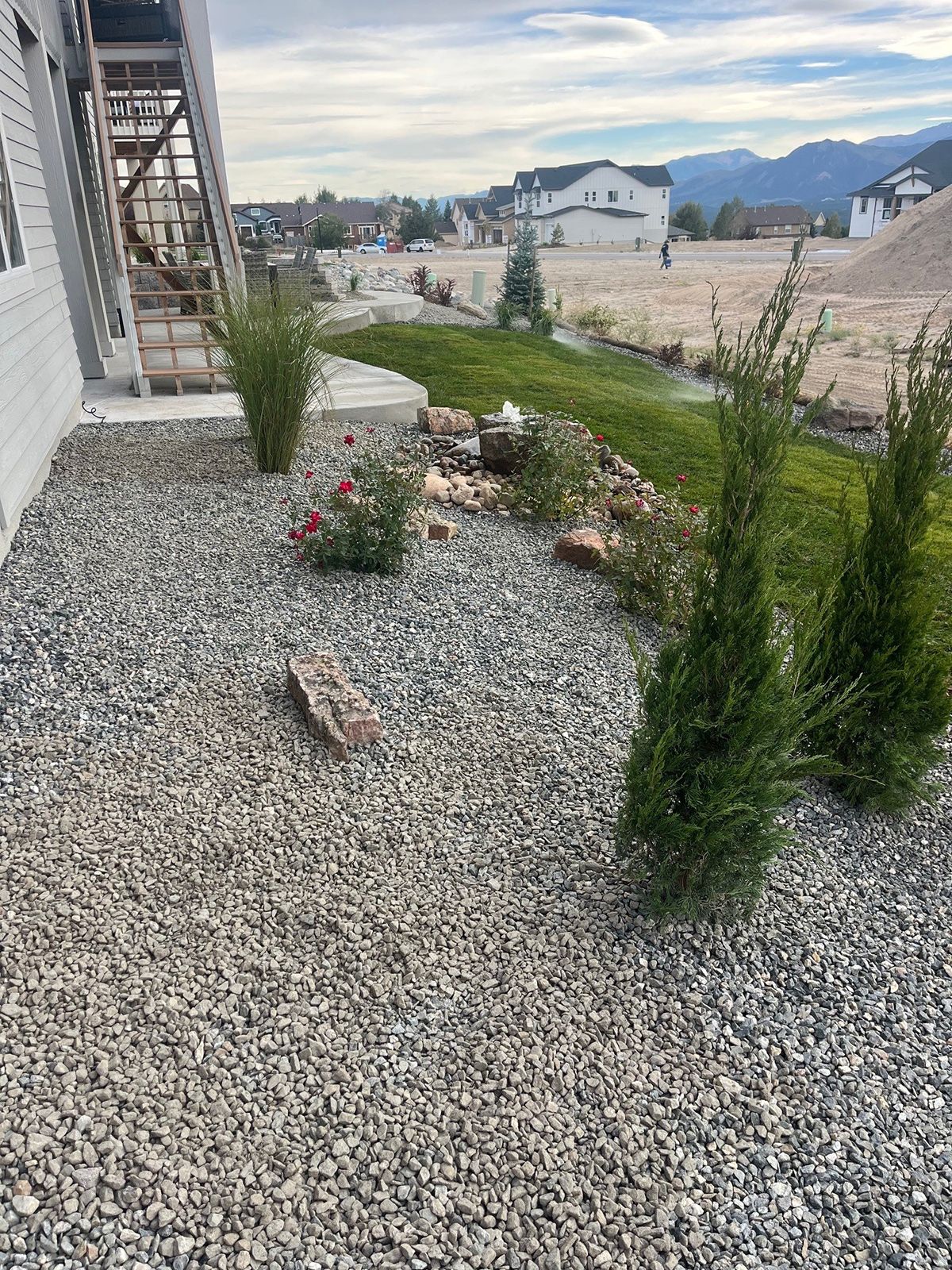 Gravel yard with green bushes, small trees, and flowers near a house under a cloudy sky.