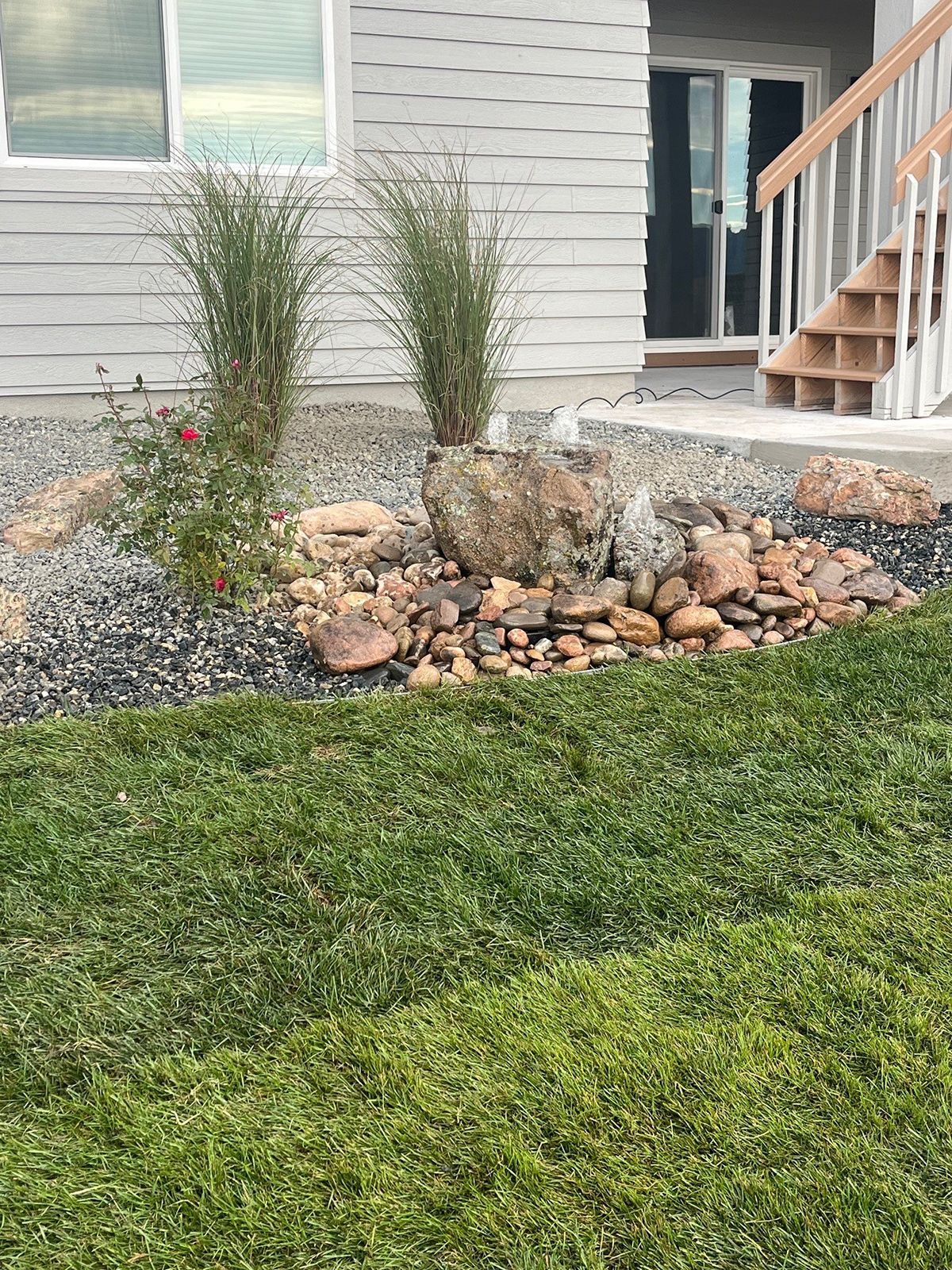 Small fountain surrounded by rocks and plants in a landscaped yard near a house.