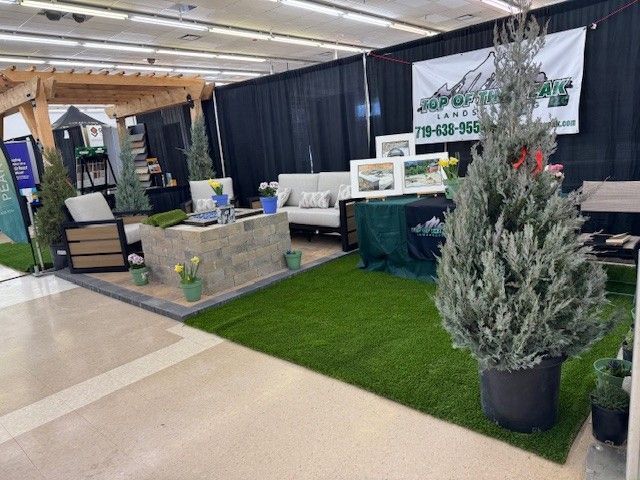 A garden landscaping booth with artificial turf, outdoor furniture, and evergreen trees. The booth's backdrop is a black curtain, and the banner says