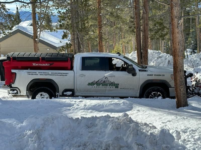 White truck with a snowplow clearing snow in a snowy wooded area. The truck has a red container on the back.