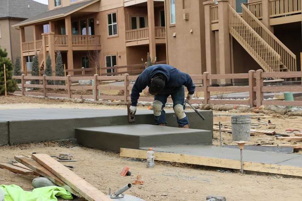 Construction worker smoothing fresh concrete on steps outdoors. He wears a blue jacket, kneepads, and is working near a brown house.