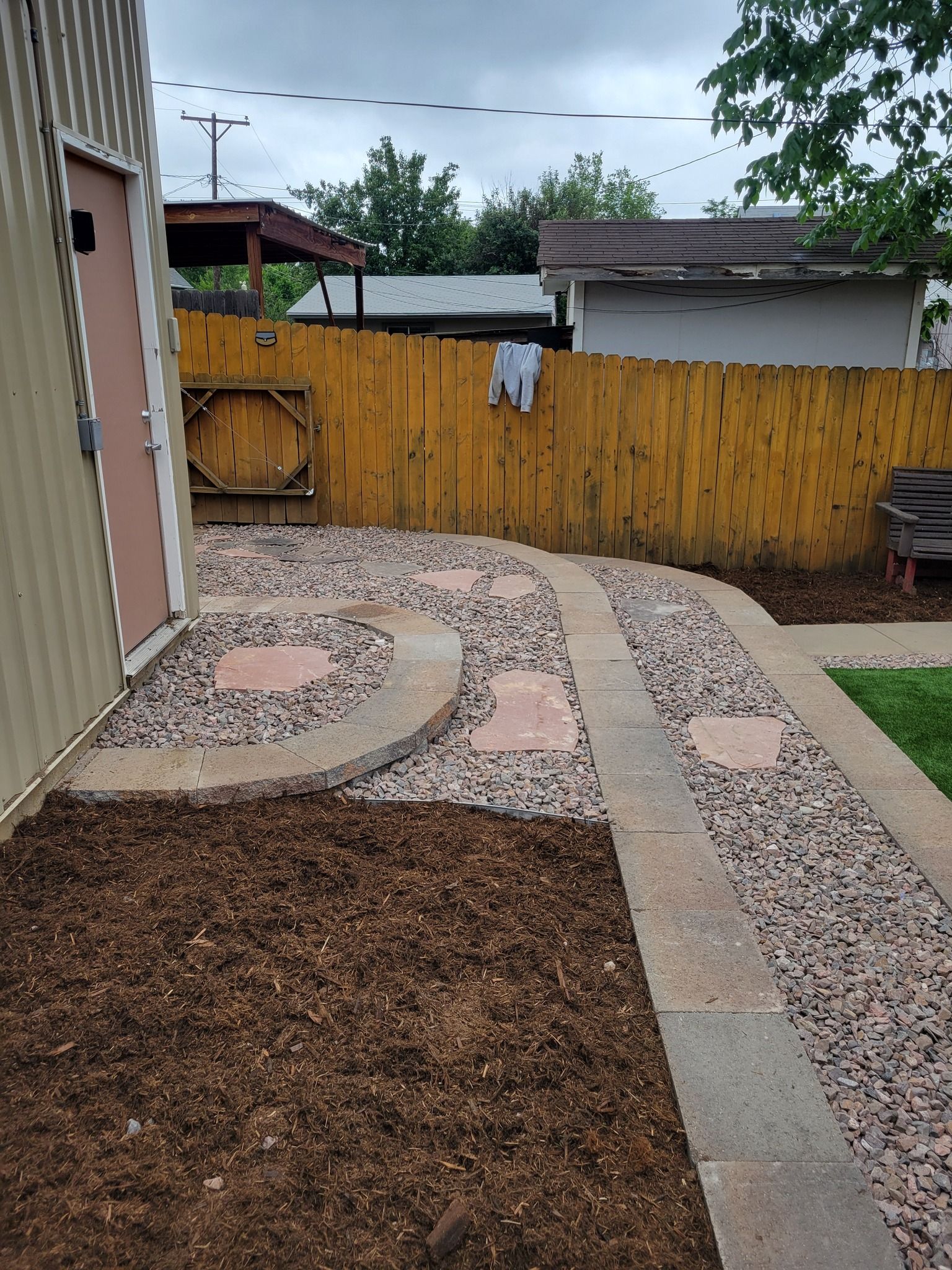 Pathway with red stone pavers and gravel, leading to a wooden fence with a shed on the left.
