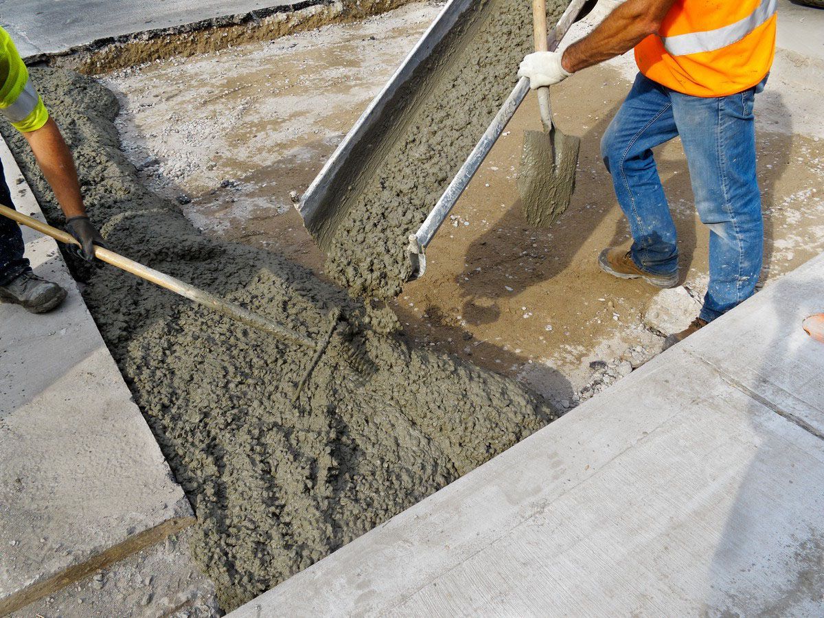 Workers pouring concrete into a prepared section of a road, one using a chute and the other with a shovel; the concrete is gray.