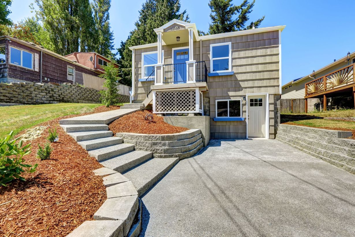 A two-story house with a concrete driveway, steps, and retaining walls. The house has tan shingles, a blue door, and a garage.