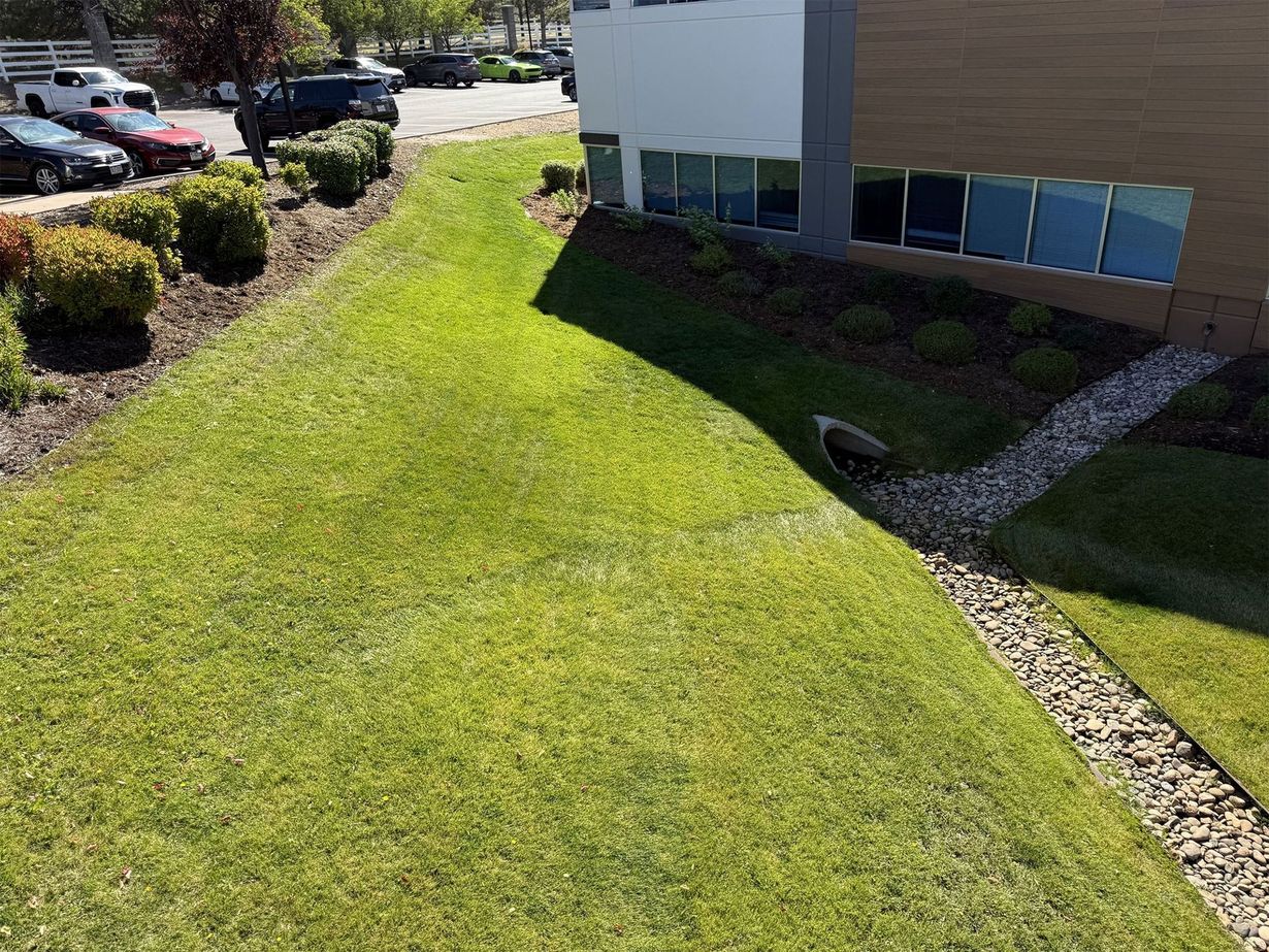 Green lawn slopes towards a gravel-lined drainage channel next to a building with tinted windows. Cars are parked in the background.