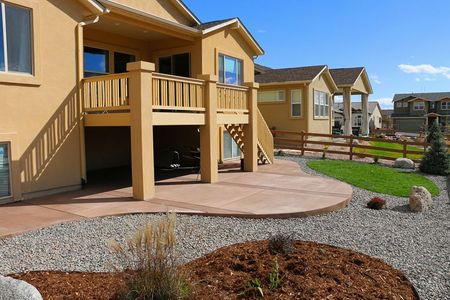 Beige house with a raised wooden deck, leading to a concrete patio surrounded by gravel and grass under a blue sky.