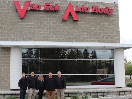 Four people standing outside Van Eck Auto Body storefront with red sign above large windows